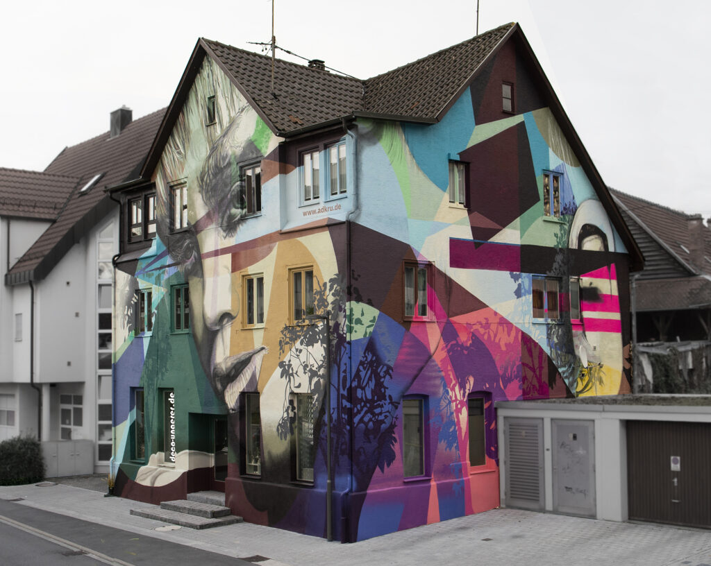 Mural Art Graffiti Haus welches die Agentur ADKRU gemalt hat. Ein bunt bemaltes Haus. Ein Portrait einer Frau, viele bunte Farben.